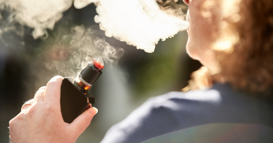 Is Vaping Bad for Your Skin? Effects on Ageing, Acne, and Complexion 1 Man holding a vape pen and inhaling, with vapor visible around him.