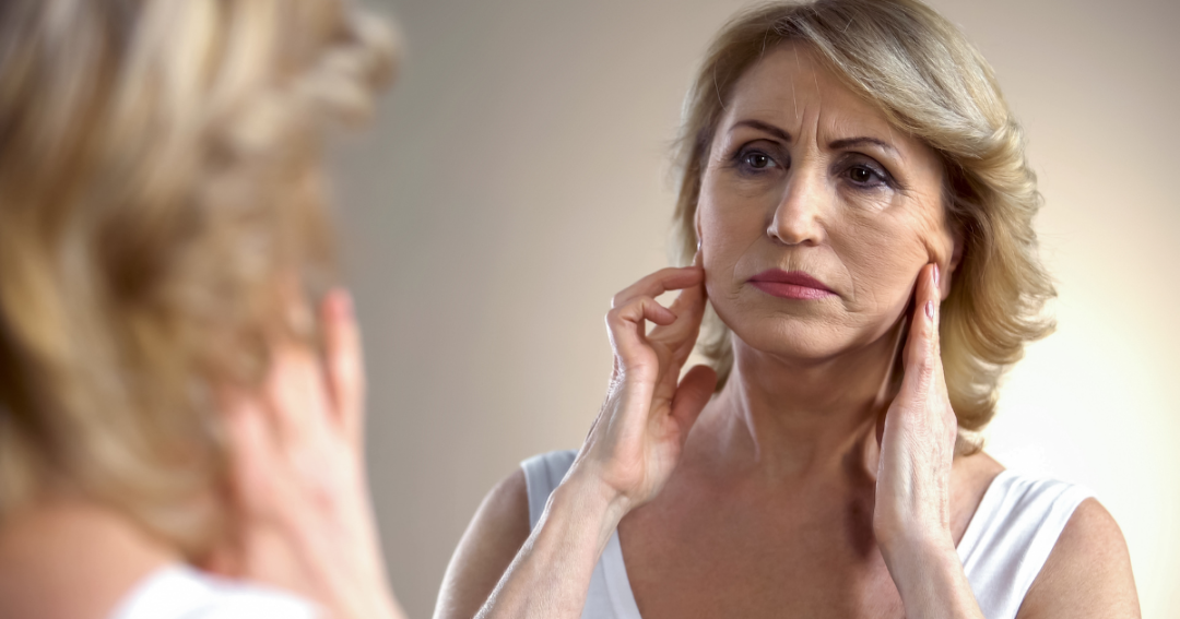 A woman examining her face closely in a mirror, assessing changes in her skin and facial volume.